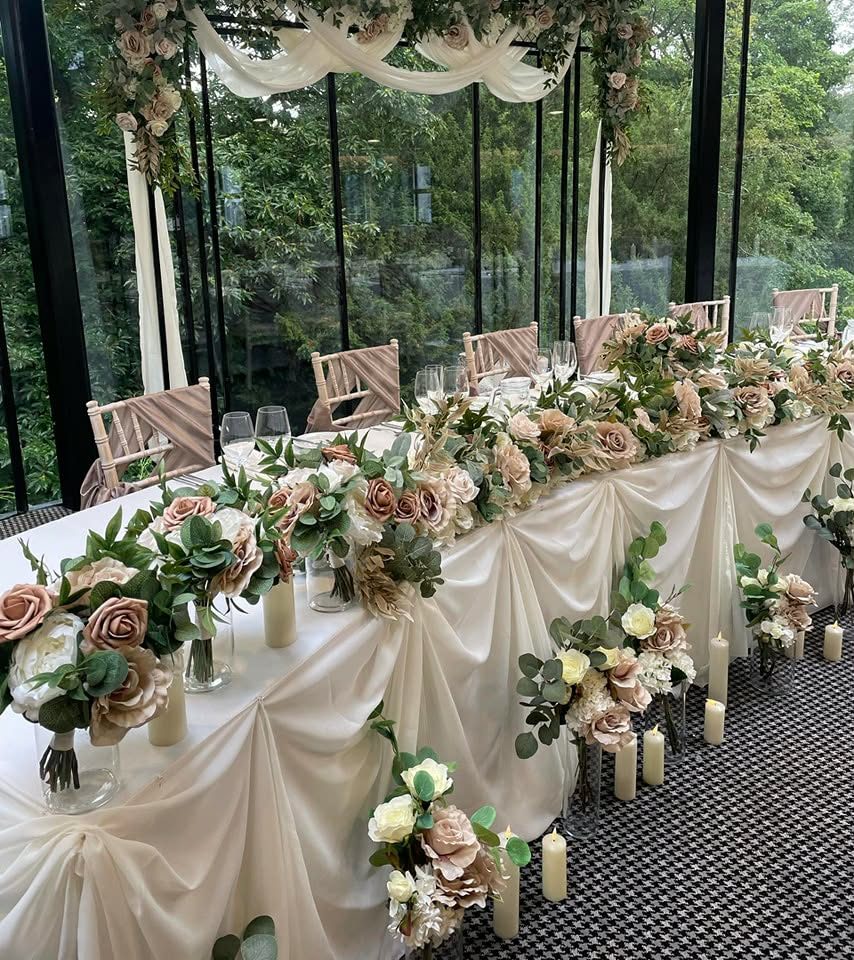 A top table in a glass-walled room, swagged in ivory satin with a garland of dusty pink and cream roses, blush chair sashes and pillar candles.