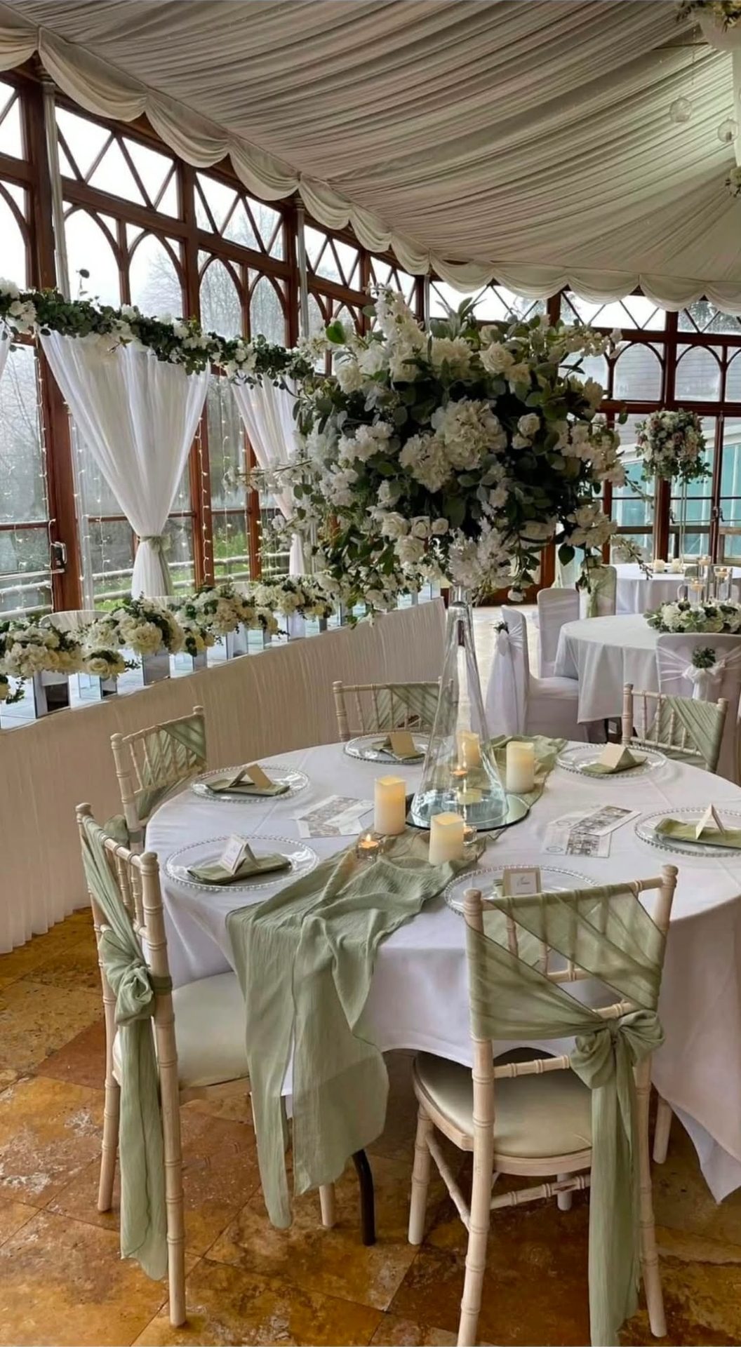 Round tables in a conservatory with sage green chair sashes and runners, glass charger plates, LED candles and a tall white floral centrepiece.