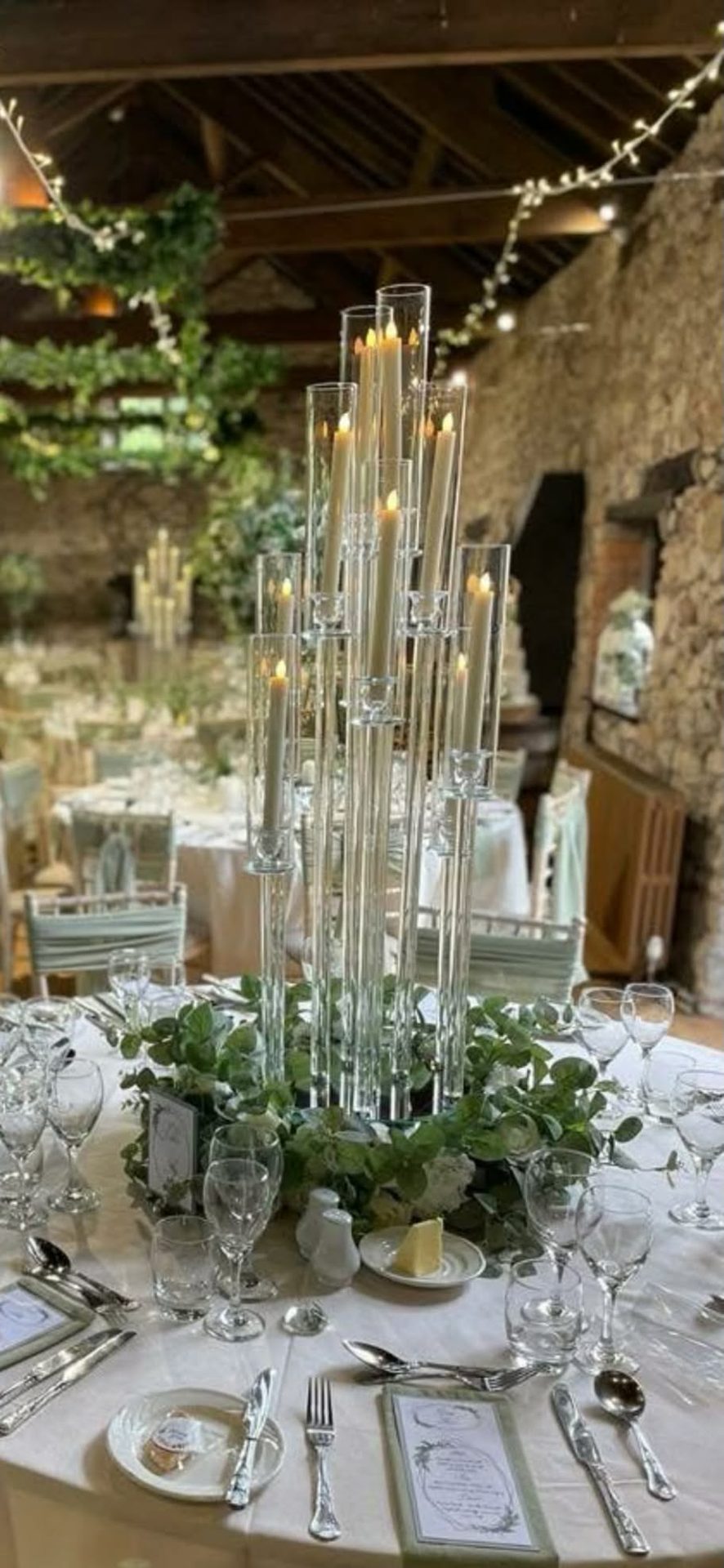 A tall glass candelabra centrepiece with taper candles in hurricane tubes, ringed with eucalyptus, in a beamed stone barn.