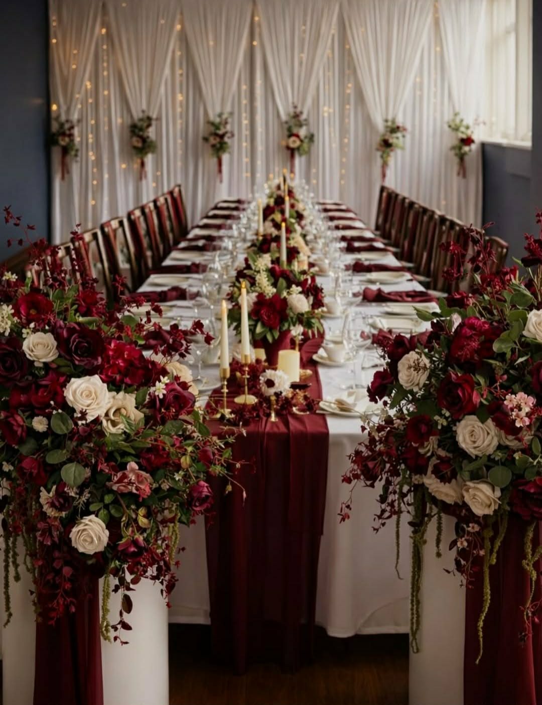 A long table dressed in burgundy and ivory - deep red rose displays on white plinths, burgundy runner and napkins, and a draped fairy-light backdrop.