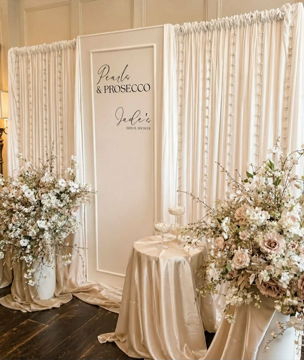 A Pearls and Prosecco bridal shower backdrop - champagne satin drapes strung with pearls, a personalised sign and neutral floral displays.